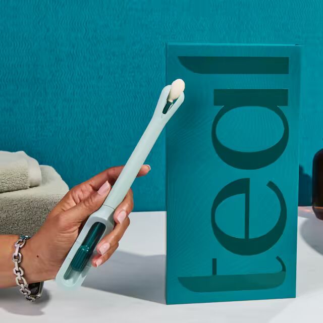 A hand holds up the light-teal colored Teal Wand self-collection device next to its vertical product packaging on a white marble bathroom vanity, showcasing its modern, user-friendly design.